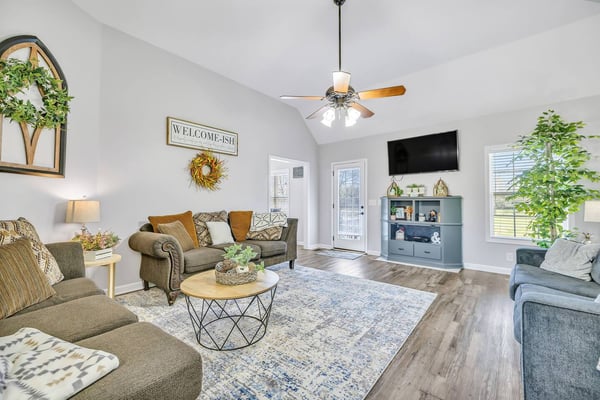 Bright open-plan living room with gray and blue upholstered seating, TV on white storage unit, ceiling fan, and wood barn door leading to home office area.